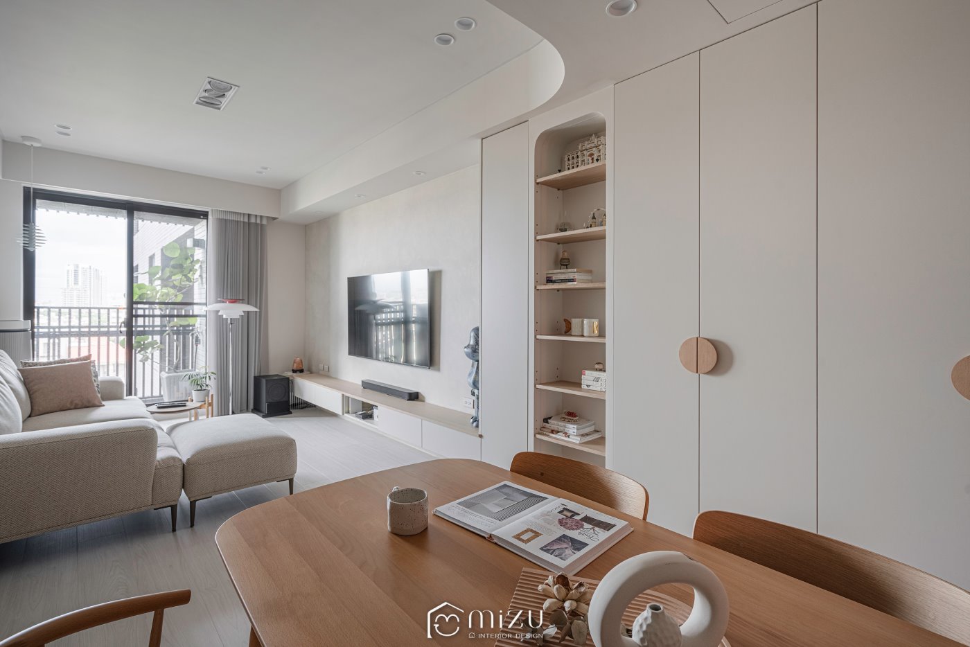 餐桌視角望向客廳，白色電視牆、弧形展示櫃與米色沙發搭配淺木紋地板 | GP7109 萊博淺橡 超耐磨木地板 | 明峻地板
